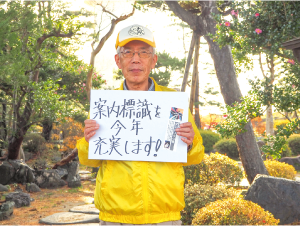 高橋さんが「案内標識を今年充実します」と書かれたボードを持った写真