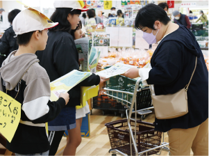 大鷲小学校4年生が西区「いっぺこーと」店内でチラシを女性客に配布をしている写真