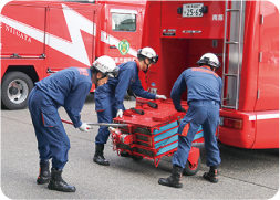 消防車後方に格納してある箱型の道具を紺色のユニホームにヘルメットを着用した３人の隊員が点検している写真